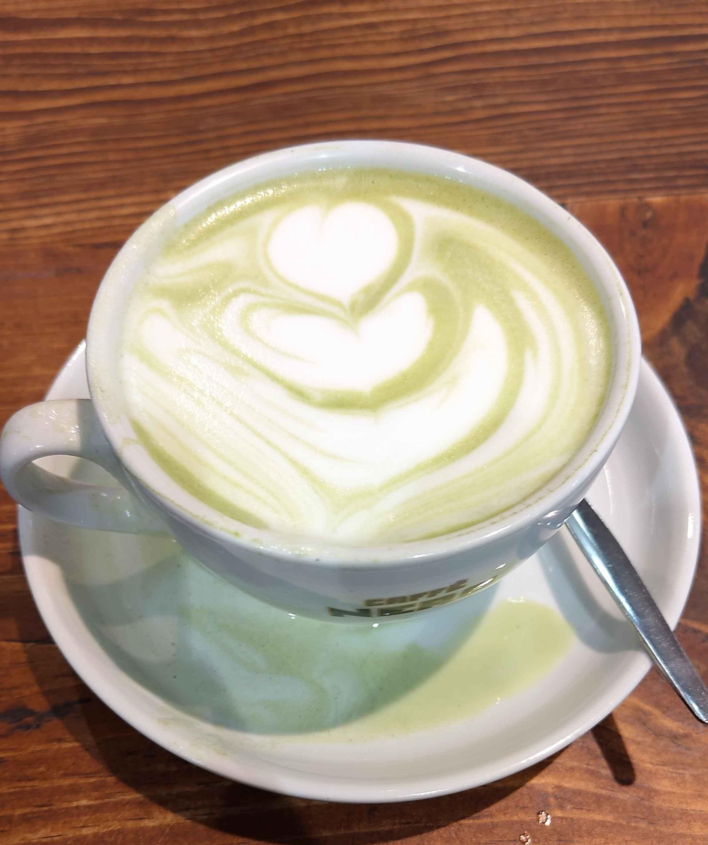 A photo of a coffee cup with a matcha latte inside. There is a foam art pattern on top.